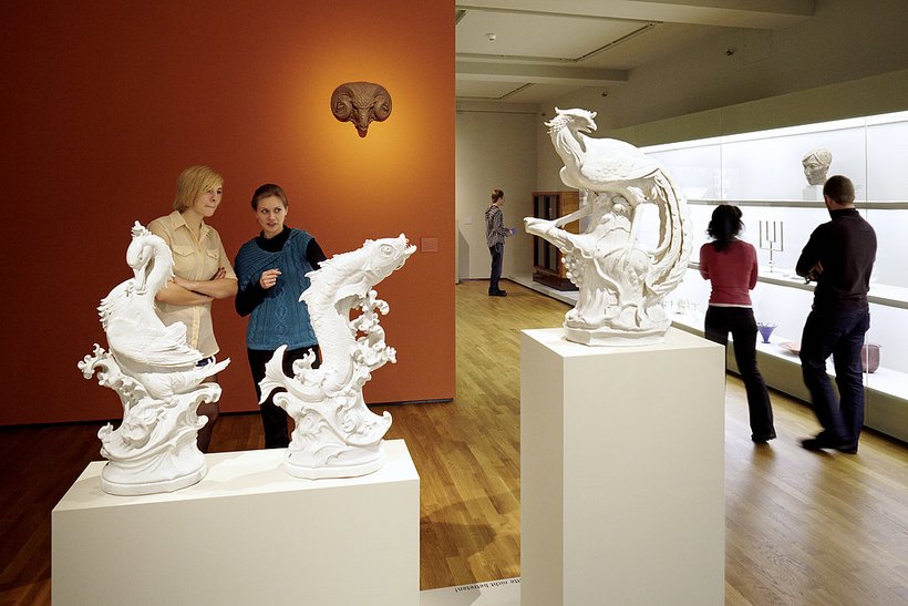 Ausstellungsansicht: links stehen zwei Personen vor einer orangenen Wand, rechts stehen zwei Personen vor einer Vitrine, im Vordergrund 3 weiße Skultpturen