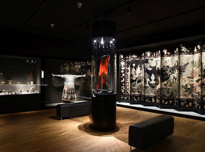 Ausstellungsansicht: abgedunkelter Raum, rechts ein asiatischer Paravent, in der Mitte eine Vitrinen mit einem roten Objekt, im Hintergrund weitere Objekte und Sitzgelegenheiten