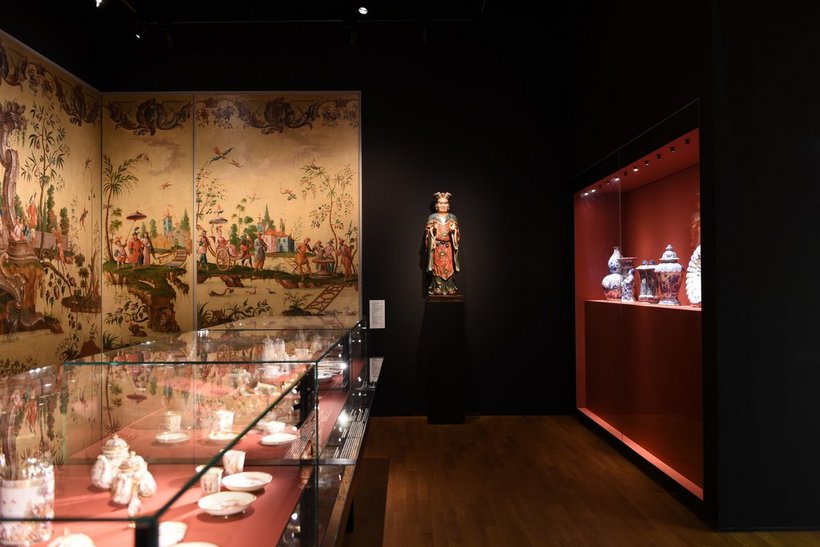 Ausstellungsansicht: abgedunkelter Raum mit einer flachen Vitrine mit Porzellan und einer Wandtapete angelehnt an chinesische Kunst, rechts eine weitere Vitrine