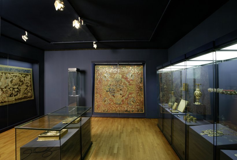 Ausstellungsansicht: dunkelblaue Wände, mehrere Vitrinen im Raumverteil, an der Wand ein großer gold-roter Teppich hinter Glas