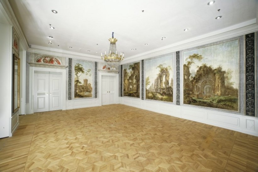 Ausstellungsansicht: ein leerer Raum mit Kronleuchter, die Wände sind mit bemalten Tapeten verziert, die bewachsene Ruinen zeigen