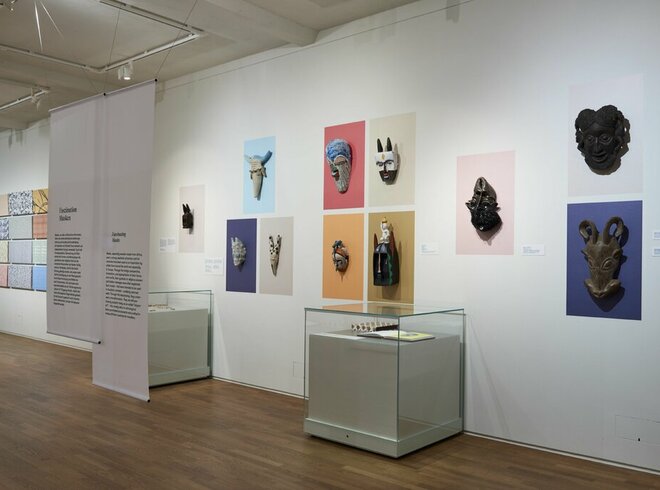 Innenansicht einer Kunstgalerie mit einer Vielfalt an ausgestellten Masken auf farbigen Hintergründen und Informationspanels an den Wänden, sowie Vitrinen mit kleineren Kunstobjekten, in einem gut beleuchteten Raum mit Holzboden