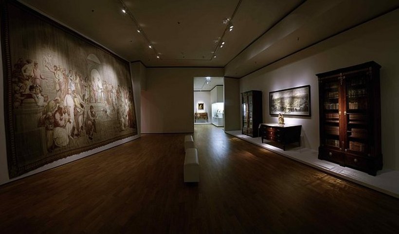 Ausstellungsansicht: abgedunkelter Raum, links ein wandfüllender Teppich, rechts verschiedene Möbelstücke