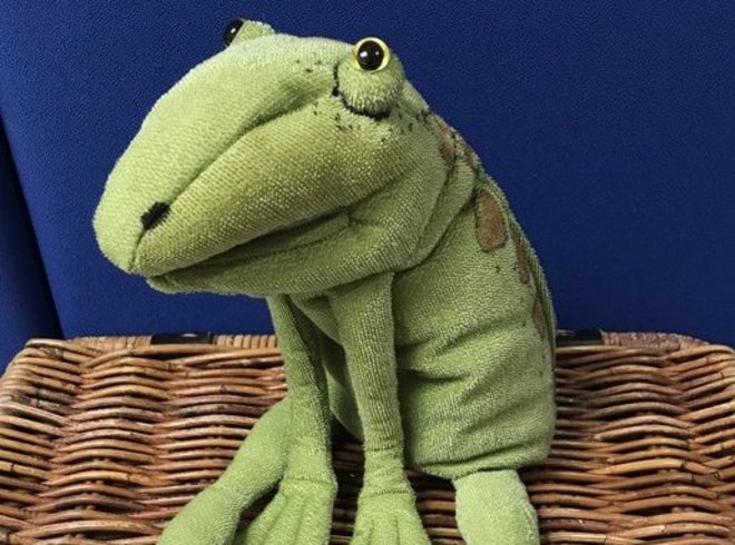 Flauschige Handspielpupe, welche einen grünen Frosch darstellt der auf einem Korb sitzt