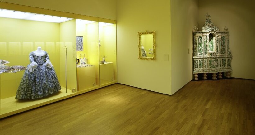 Ausstellungsansicht: gelbe Wände, links eine große Vitrine mit einem historischen Abendkleid, rechts hinten ein reich verzierter Kachelofen