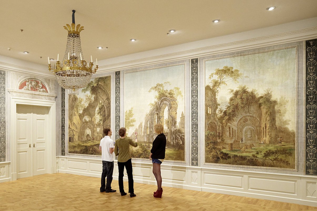 Drei Personen stehen vor einer gemalten Tapete, auf der verfalle und bemalte Architektur zu sehen ist