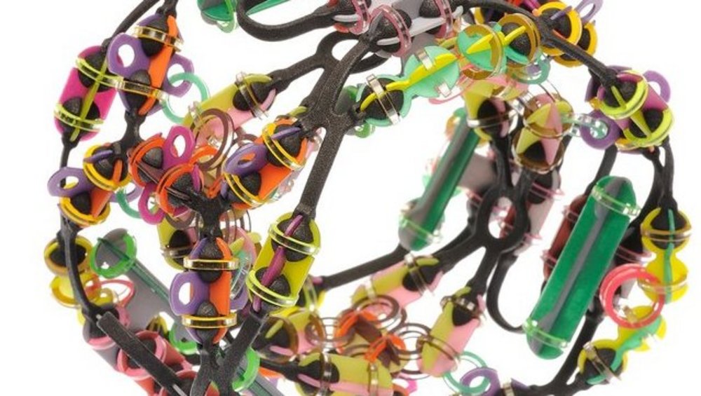 künstlerisches Objekt bestehend aus verschlungenen bunten Gummibändern und mehreren ineinander verhakten Scherengriffen, die eine abstrakte, kugelförmige Skulptur bilden, auf einem weißen Hintergrund