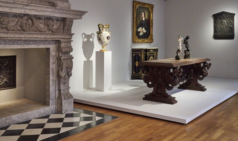 Ausstellungsansicht: links ein großer Kamin aus Stein, zentral steht ein Tisch aus dunklem Holz, darum eine Vasen, Skulpturen und Möbel