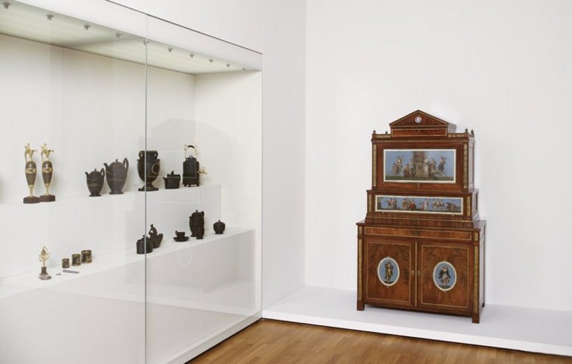 Ausstellungsansicht: links eine große Vitrine mit Gefäßen, rechts ein Möbelstück aus Holz