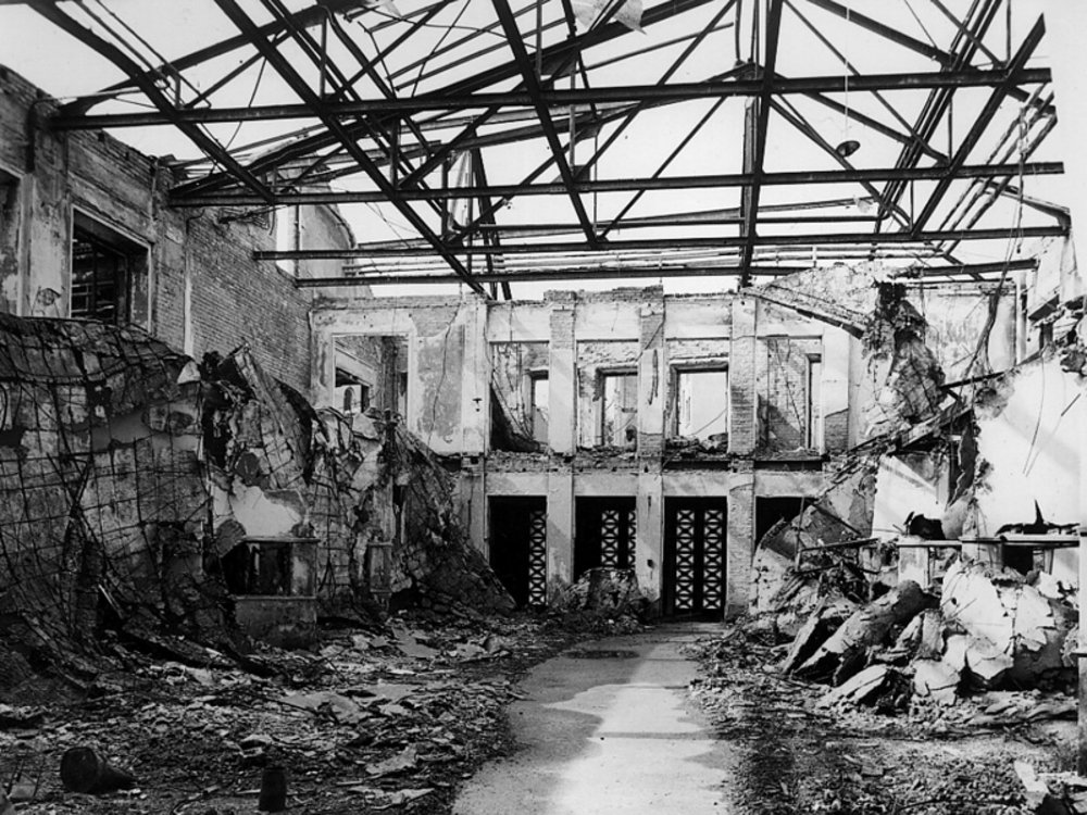 schwarz-weiß Fotografie des zerstörten Gebäudes, Dach fehlt, nu die Stahlkonstruktion ist zu sehen, links und rechts Schuttberge