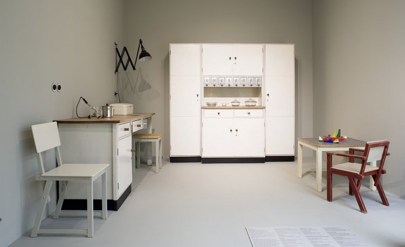 Ausstellungsansicht: in der Mitte ein weißes Küchenbufett, links eine Küchenzeile und ein Stuhl, rechts Kindermöbbel