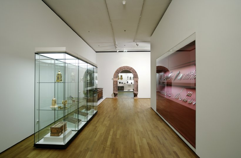 Ausstellungsansicht: links eine Vitrine im Raum, rechts eine verglaste rote Wand, im Hintergrund ein Torbogen