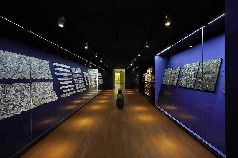 Ausstellungsansicht: abgedunkelter Raum, mit dunkelblauen Wänden, links und rechts wandhohe Vitrinen mit textiler Spitze