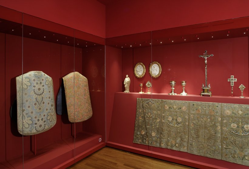 Ausstellungsansicht: rote Wände, eine große Vitrine über Eck zeigt religiöse Gegenstände und Textilien