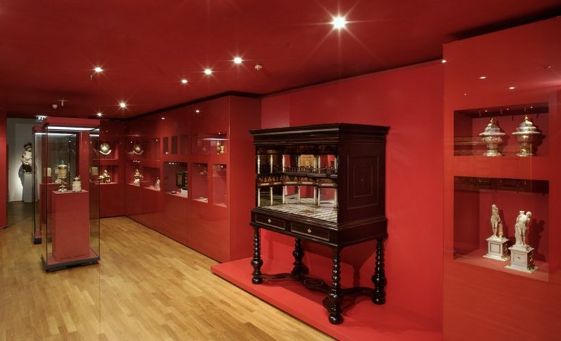 Ausstellungsansicht: rote Wände, viele kleine Vitrinen in der Wand eingelassen und Möbelstück aus dunklem Holz
