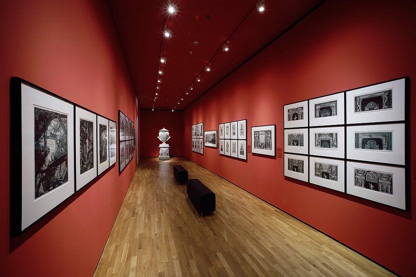 Ausstellungsansicht: rote Wände, links und rechts an den Wänden gerahmte Grafiken