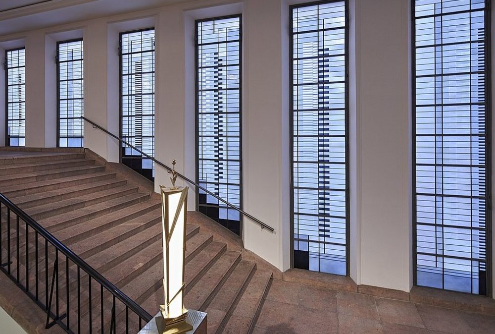 Treppenhaus, im Hintergrund eine Reihe länglicher Buntglasfenster, im Vordergrund eine goldene Leuchte auf dem Treppenansatz