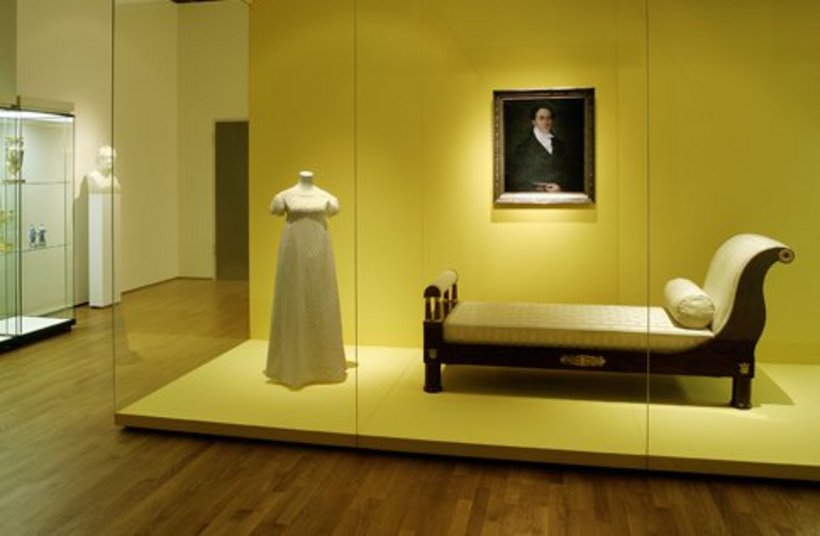 Ausstellungsansicht: eine wandhohe Vitrine mit gelber Wand, darin ein Abendkleid, eine Liege und ein Portrait
