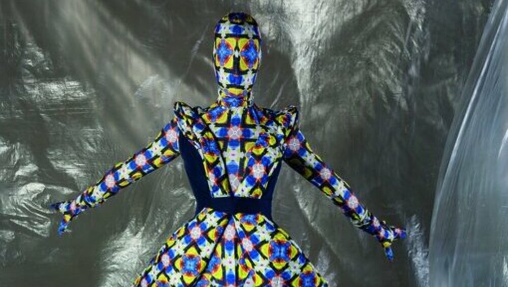 Eine Modepuppe in einem vollständig bedeckenden Kleid mit buntem grafischem Muster steht vor einem silbernen Metallic-Hintergrund