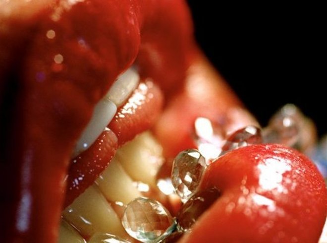 Bild von einem halb geöffneten Mund mit einer Nahaufnahme der Lippen und Zähne. Eine Kette mit Kristallen wird von der Unterlippe gehalten