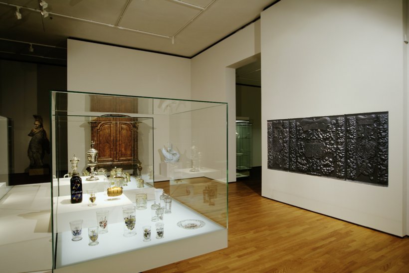 Ausstellungsansicht: links eine große Vitrine im Raum, recht an der Wand ein Relief, im Hintergrund ein Holzschrank