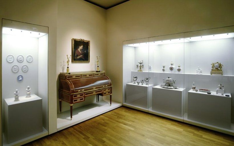 Ausstellungsansicht: rechts eine große Vitrine, links eine kleine Vitrine mit Porzellan, dazwischen ein Sekretär aus Holz