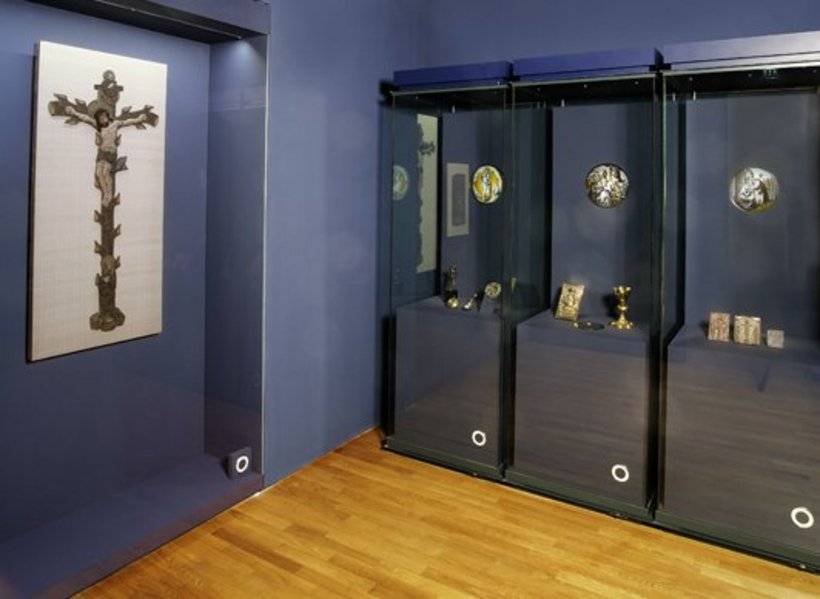 Ausstellungsansicht: dunkelblaue Wände, links eine Vitrine mit Jesus am Kreuz, rechts drei Vitrinen mit religiöser Kunst