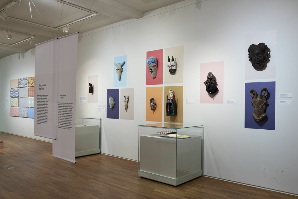 Innenansicht einer Kunstgalerie mit einer Vielfalt an ausgestellten Masken auf farbigen Hintergründen und Informationspanels an den Wänden, sowie Vitrinen mit kleineren Kunstobjekten, in einem gut beleuchteten Raum mit Holzboden